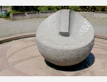 Steinerne Sonnenuhr in Kugelform auf einem Platz mit gepflastertem Boden und umliegender Vegetation; auf der Sonnenuhr ist der Schriftzug „Sei dir deiner Zeit bewusst“ erkennbar.