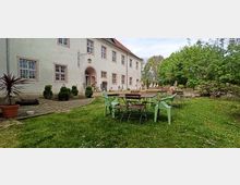 Hinterhof eines historischen Gebäudes mit hell verputzter Fassade und mehreren kleinen Fenstern mit dekorativem Gitter. Im Vordergrund stehen Gartenmöbel auf einer Wiese, umgeben von Bäumen und Sträuchern.