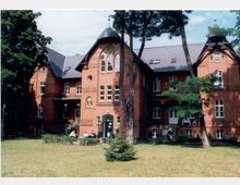 Historisches Backsteingebäude mit spitzem Giebeldach und gotischen Fensterelementen, umgeben von Bäumen und einem gepflegten Rasen. Im Vordergrund sind mehrere Personen und ein kleiner Gartenbereich zu sehen.