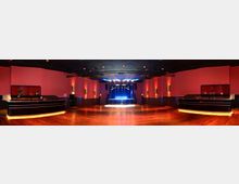 Großer Veranstaltungsraum mit Holzfußboden, einer Bühne mit blauen Lichtern im Hintergrund und symmetrisch angeordneten Wandleuchten an den roten Wänden. Seitlich befinden sich zwei schwarze Tresen mit indirekter Beleuchtung.