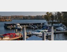 Jachthafen an einem See mit festgemachten Motorbooten und kleinen Hausbooten, umgeben von herbstlichen Bäumen und einer ruhigen Wasserfläche. Im Hintergrund sind Windräder und vereinzelte Gebäude sichtbar.