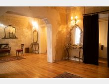 Ein Innenraum mit abgeblätterten Wänden und Holzboden, ausgestattet mit antiken Möbeln, darunter ein vergoldeter Konsolentisch mit einem verzierten Spiegel und ein Holzstuhl mit rotem Polster. Der Raum ist durch warmes Licht beleuchtet, und ein schwarzer Vorhang trennt einen Eingang rechts im Bild.