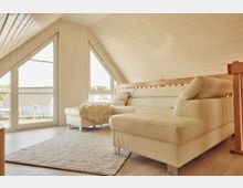 Wohnzimmer in einem Dachgeschoss mit schräger Holzdecke, großen dreieckigen Fenstern und einem beigen Ledersofa. Der Raum ist hell, mit Holzfußboden und einem Teppich, sowie Blick auf mehrere Häuser im Freien.