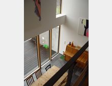 Innenraum eines modernen Wohnraums mit hohen Decken, großen bodentiefen Fenstern und einer klaren, minimalistischen Einrichtung aus Holz. Zu sehen sind ein Esstisch mit Stühlen, ein kleines Klavier oder Sideboard, sowie ein angrenzender Außenbereich mit Holzterrasse.