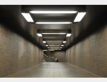 Unterirdischer Fußgängertunnel mit braunen Ziegelwänden, einem gefliesten Boden und einer Reihe von rechteckigen Deckenleuchten. Am Ende des Tunnels ist eine Rolltreppe sichtbar.