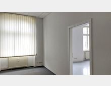 Heller Büroraum mit weißen Wänden, einem großen Fenster mit vertikalen Lamellenjalousien und einem Heizkörper darunter; durch eine offene Tür ist ein angrenzender Raum mit ähnlich gestaltetem Fenster und weißem Boden sichtbar.