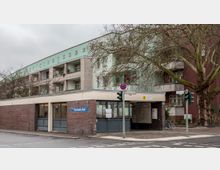 Eingang zu einer U-Bahn-Station namens "Parchimer Allee" mit einem flachen Gebäude aus Backstein und Glas, darüber ein mehrstöckiges Wohngebäude mit Balkonen. Im Vordergrund stehen ein großer Baum und eine Fußgängerampel an einer Straßenkreuzung.