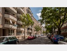 Wohnstraße mit mehrstöckigen Wohngebäuden auf der linken Seite, die über Balkone verfügen. Rechts und links der Straße stehen parkende Autos, und Bäume säumen den Gehweg, die Schatten auf die Fahrbahn werfen.