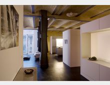 Wohnraum in einem modernen Loft mit sichtbarem Gewölbedecken aus gelben Ziegeln und Stahlträgern. Der Raum ist lichtdurchflutet, zeigt minimalistische Möbel und verbindet Wohn- und Küchenbereich; im Hintergrund sind große Fenster mit Blick nach außen.