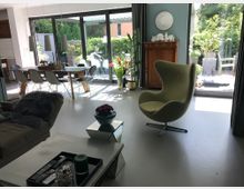 Helles modernes Wohnzimmer mit großen Glas-Schiebetüren, die zu einem Gartenbereich führen. Im Raum stehen ein grüner Sessel, ein Esstisch mit Stühlen und eine Vitrine, umgeben von Pflanzen und Dekoration.