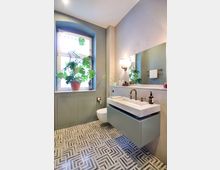 Modernes Badezimmer mit geometrisch gemustertem Boden, einem großen Spiegel, einem Waschbecken aus Marmor mit grünem Unterschrank und goldenen Armaturen. Ein Fenster mit Pflanzen in Terrakottatöpfen bringt Licht in den Raum, der in neutralen Farbtönen gehalten ist.