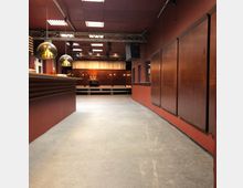 Ein Veranstaltungsraum mit einer Holztheke auf der linken Seite und geschlossenen Holztüren auf der rechten Seite. Im Hintergrund befindet sich eine erhöhte Bühne mit Vorhängen und Beleuchtung, umgeben von einer roten Wandgestaltung und moderner Deckenkonstruktion.