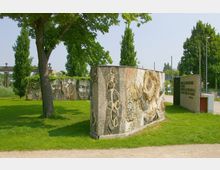 Eine kleine Grünfläche mit Bäumen, auf der künstlerisch gestaltete Mosaikwände stehen. Die Wände sind mit abstrakten Mustern und Porträtdarstellungen verziert, und eine Steintafel mit Text ist ebenfalls sichtbar.