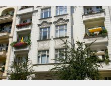 Fassade eines historischen Wohngebäudes mit hellen Wänden, dekorativen Stuckelementen und Balkonen, die mit Blumen und Pflanzen geschmückt sind. Im Vordergrund steht ein Baum, der teilweise das Gebäude verdeckt.