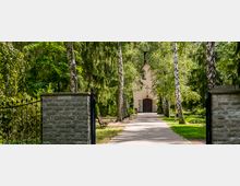 Pflasterweg, gesäumt von hohen Birken und Tannen, führt zu einer kleinen Kapelle mit cremefarbener Fassade und Holztür. Die Umgebung ist von dichtem Grün und einer Ziegelsteinmauer mit einem schmiedeeisernen Tor geprägt.