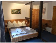 Schlafzimmer mit dunklem Holzbalken, einem Doppelbett mit heller Bettwäsche und dekorativen Kissen, eine Holzkommode, ein Korbstuhl sowie ein gerahmtes Bild an der Wand. Die Wände und Möbel sind in warmem Holzton gehalten.