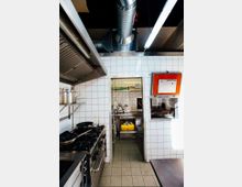 Kleine gewerbliche Küche mit gefliesten weißen Wänden und grauem Boden. Sichtbar sind ein Gasherd mit mehreren Pfannen, eine Durchreiche mit einer roten Klappe, ein Belüftungssystem an der Decke und eine angrenzende Lagerecke mit Küchenutensilien.