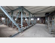 Industrieähnlicher Dachbodenraum mit freiliegenden Stahlträgern und Holzboden, umgeben von unverputzten Ziegelwänden. Im Raum befinden sich Metalltreppen, Geländer, mehrere Fenster und eine Tür an der Rückwand.