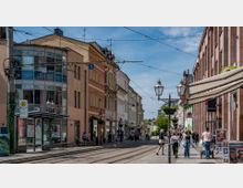 Straßenszene in einer belebten Innenstadt mit Straßenbahnschienen, Fußgängern, Geschäften und historischen Gebäuden mit verzierten Fassaden. Im Vordergrund ist eine Straßenbahnhaltestelle mit einem Wartehäuschen und einer Laterne zu sehen, während Markisen und Schilder die Geschäfte säumen.