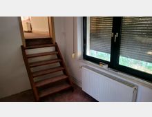Ein kleiner Innenraum mit einer Holztreppe, die zu einem höher gelegenen Bereich führt. Rechts im Bild befindet sich ein großes Fenster mit geschlossenen Rollläden und darunter ein weißer Heizkörper.