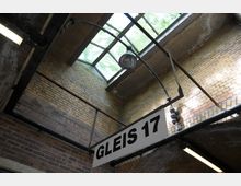 Ansicht eines historischen Bahnhofsbereichs mit gelben und roten Backsteinwänden, darüber ein verglastes Dach mit Metallstreben. Im Vordergrund hängt ein Schild mit der Aufschrift "Gleis 17".