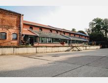 Backsteinbau im Industriestil mit Satteldach und großer Glasfront, flankiert von Betonfundamenten. Im Vordergrund ein leerer, geteilter Hofbereich, im Hintergrund Bäume.