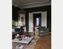 Wohnzimmer mit dunkelgrünen Wänden, einem klassischen weißen Kamin mit Verzierungen, einer Doppeltür, die zu einem Flur führt, und Holzboden. Graue Sofas, ein niedriger Couchtisch mit Büchern und Dekorationen sowie ein gemusterter Teppich bestimmen die Einrichtung.