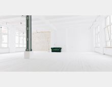 Weiß gestrichener, leerer Raum mit sichtbarer Backsteinwand, großen Fenstern auf beiden Seiten und einem grünen, klassischen Sofa in einer Ecke; zentrale Metallstütze und frei hängende Kabel sichtbar.