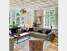 Wohnzimmer mit großen Fenstern, die den Raum mit Tageslicht erhellen. Eine graue und eine blaue Couch stehen um einen rechteckigen Holztisch, daneben befinden sich Zimmerpflanzen, eine Tischlampe und ein Gemälde an der Wand; die Decke hat ein dekoratives Kassettenmuster.