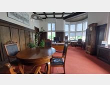 Historischer Büroraum mit holzvertäfelten Wänden, einem runden Holztisch mit lederbezogenen Stühlen im Vordergrund, einem Schreibtisch mit Computer vor großen Fensterfronten im Hintergrund, und einer Standuhr an der rechten Seite. Der Raum ist mit rotem Teppichboden ausgelegt und hat dekorative Deckenpaneele.