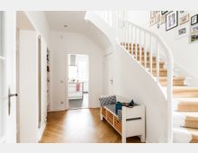 Heller Eingangsbereich eines Hauses mit geschwungener weißer Treppe und Teppichstufen, Holzboden in Fischgrätmuster und einer Sitzbank mit Stauraum. Im Hintergrund ist eine offene Tür, die in eine helle Küche führt, sowie an der Wand gerahmte Fotos.