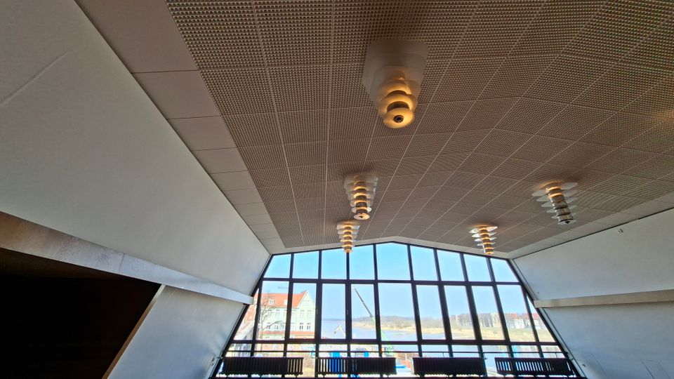 Innenraum einer modernen Halle mit hoher, perforierter Decke und mehreren hängenden Leuchten. Eine große, bogenförmige Fensterfront mit schwarzem Rahmen blickt auf Wasser, Uferlandschaft und Gebäude im Hintergrund.