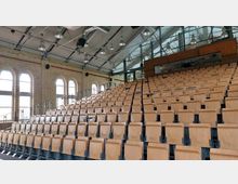 Ein moderner Hörsaal mit ansteigenden Sitzreihen aus hellem Holz und integrierten Klappsitzen. Im Hintergrund sind gemauerte Wände mit hohen, bogenförmigen Fenstern sowie eine verglaste Rückwand sichtbar, die Tageslicht in den Raum lässt.