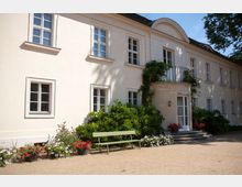 Helles, zweistöckiges Gebäude mit weißer Fassade, Sprossenfenstern und einem kleinen, halbkreisförmigen Balkon über einer Tür mit Glasflächen. Vorgarten mit Blumen in Beeten und Töpfen sowie einer grünen Bank, umgeben von Büschen.