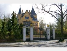 Gelbes Herrenhaus im historischen Stil mit mehreren Türmen und spitzen Dächern, umgeben von einem schmiedeeisernen Zaun und Bäumen. Im Vordergrund ein großes Tor mit Steinsäulen und Laternen, dahinter ein gepflegter Garten.