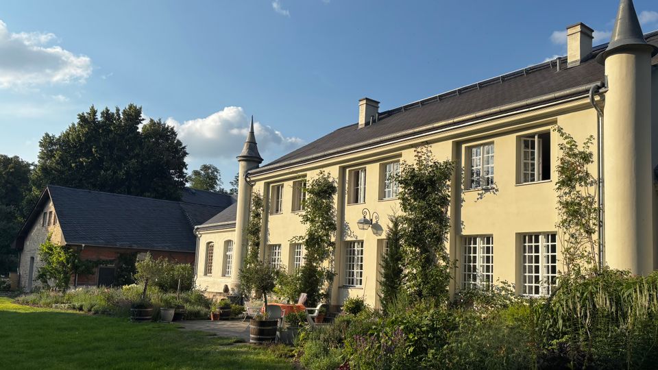 Zweistöckiges, helles Landhaus mit dunklem Satteldach und turmartigen Eckpfeilern mit Spitzdächern, davor eine Terrasse und ein bepflanzter Garten. Links steht ein Nebengebäude mit Schieferdach; die Anlage ist von Rasenflächen und Bäumen unter blauem Himmel umgeben.