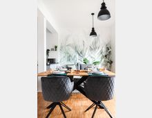 Essbereich in einer modernen Wohnung mit Holzboden. Ein Esstisch aus Holz mit gedeckten Plätzen steht vor einer Wand mit tropischem Blättermuster, eingerahmt von zwei grauen, gepolsterten Stühlen und schwarzen Hängeleuchten.