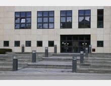 Moderne Gebäudefassade mit beigefarbenen Steinplatten und großen, dunklen Fenstern. Vor dem Eingang führen breite Treppenstufen mit niedrigen Laternen zu einer Glas-Doppeltür, rechts daneben ist die Hausnummer 62 angebracht.