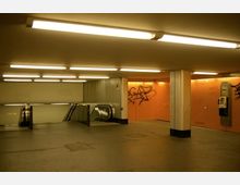 Ein U-Bahn-Eingangsbereich mit hell beleuchteten Decken, orangefarbenen Wänden teilweise bedeckt mit Graffiti, Rolltreppen auf der linken Seite und Wandtelefonen auf der rechten Seite. Der Boden des Bereichs ist schlicht und grau gehalten.