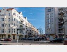 Straßenszene in einer Wohngegend mit mehrstöckigen Altbau-Mietshäusern. Die Gebäude haben helle Fassaden, Balkone und rote Dächer; im Erdgeschoss befinden sich Geschäfte. Straßenschilder zeigen die Kreuzung der Apostel-Paulus-Straße und Eisenacher Straße. Autos sind entlang der Straße geparkt.