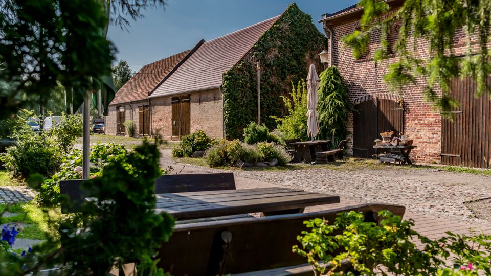 Innenhof eines ländlichen Bauernhofs mit Kopfsteinpflaster, umgeben von Backstein- und Scheunengebäuden mit Holztoren und Satteldächern. Im Vordergrund stehen Holzbänke und Tische in einem begrünten Gartenbereich mit Sträuchern und Bäumen.