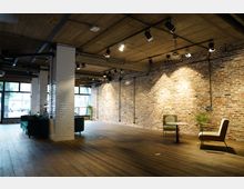 Geräumiger Innenraum im Industriestil mit freiliegenden Ziegelwänden, Holzboden und einer Betondecke mit schwarzen Schienenlichtern. Der Raum ist möbliert mit grünen Sofas, Stühlen und einem kleinen Tisch, und große Fenster lassen Tageslicht herein.