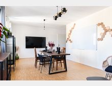 Modern eingerichteter Raum mit einem rechteckigen Esstisch aus Holz, umgeben von verschiedenen Stühlen. An der Wand befinden sich ein großer Flachbildfernseher, eine weiße Tafel und dekorative Holzpaneele in Wabenform.