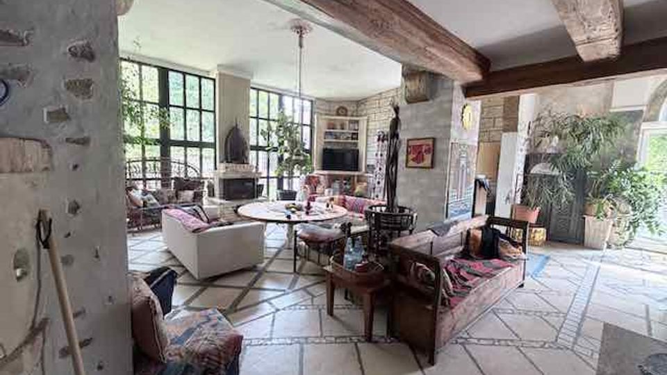 Großzügiger Wohnbereich im rustikalen Stil mit sichtbaren Holzbalken, Steinwänden und hellem Natursteinboden in geometrischem Muster. Große Fensterfronten, ein Kamin und mehrere Sofas und Sitzgruppen prägen den offenen Raum mit Blick ins Grüne.