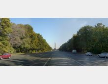 Weitläufige mehrspurige Straße, gesäumt von dicht bewachsenen Bäumen auf beiden Seiten, mit der Siegessäule als zentralem Monument in der Ferne. Am Straßenrand parken vereinzelt Autos, und der Himmel ist wolkenlos.