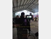 Innenansicht einer überdachten Halle mit sichtbaren Metallträgern, in der eine Filmszene gedreht wird. Im Vordergrund steht ein Kameramann mit einem Monitor, während im Hintergrund weitere Personen und Requisiten zu sehen sind, umgeben von rauchigen Lichteffekten. Auf dem Monitor ist eine Nahaufnahme von Schauspielern erkennbar.