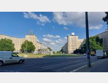 Straßenszene mit breiter Fahrbahn und Grünanlage in der Mitte, flankiert von zwei mehrstöckigen, historischen Wohngebäuden im sozialistischen Baustil. In der Mitte der Grünanlage befindet sich ein Springbrunnen, umgeben von Bäumen und Fußwegen.