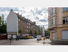Straßenszene in einem Wohngebiet mit mehrstöckigen Häusern, darunter ein gelb-beiges Eckgebäude mit Ladenfassade ("HMEHPRESS") und ein langes Ziegelwohnhaus. Autos und ein gelber Lieferwagen sind am Straßenrand geparkt, eine Radfahrerin fährt vorbei, und Bäume säumen die Straße.