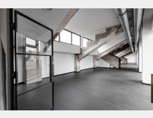 Industrieartiger Innenraum mit großen, schrägen Betonstützen, weißen Wänden und dunklem Bodenbelag. Oben verlaufen sichtbare Lüftungsrohre, und die Fensterfront lässt Tageslicht in den Raum.