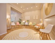 Ein moderner Warteraum mit einer Reihe bunter Stühle in Pastelltönen entlang einer beige gestrichenen Wand. Ein runder Teppich mit gelbem Musterdruck, eine Stehlampe und verschiedene Wanddekorationen sowie Pflanzen verleihen dem Raum eine einladende Atmosphäre.